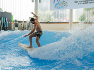 Ein Mann surft auf der stehenden Welle in einer Indoor-Anlage der Jochen Schweizer Arena. Er trägt eine Badehose und balanciert auf seinem Surfbrett, während Wasser um ihn herum spritzt.