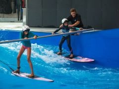2 Kinder surfen mit Hilfe einer Stange auf der stehenden Welle mit einem Instruktor