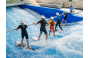Thumbnail5 Kinder surfen auf der stehenden Welle der Jochen Schweizer Arena, während die Surflehrerin ihnen erklärt, wie sie auf der Welle ohne Haltestange halten können.