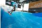 ThumbnailKinder-Surferlebnis in der Jochen Schweizer Arena: Ein Mädchen surft mit Helm und Neoprenanzug sicher auf der Indoor-Welle vor der imposanten Markenwand der Arena.