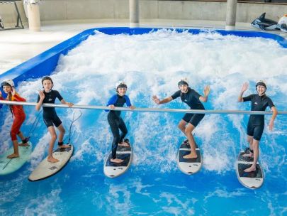 Indoor-Surfen für Kinder in der Jochen Schweizer Arena: Sechs junge Teilnehmer stehen lachend mit Surfboards auf der stehenden Welle und halten sich an einer Trainingsstange fest.