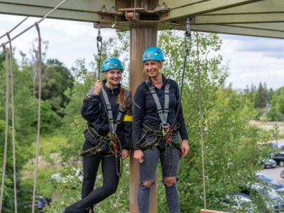 Outdoor-Kletterabenteuer in der Jochen Schweizer Arena: Zwei Frauen mit Helm und Sicherheitsausrüstung stehen lachend auf der Plattform des Hochseilgartens.