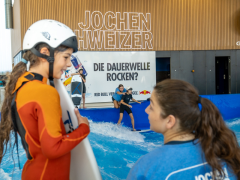 Ein Mädchen und eine Surflehrerin sind im Gespräch, während eine weitere Surflehrerin einem anderen Mädchen beim Einstieg auf die stehende Welle hilft