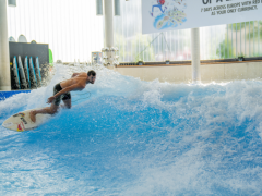 Ein Mann surft die Indoor Welle in der Jochen Schweizer Arena. Er lehnt sich in die Welle hinein und das Wasser spritzt