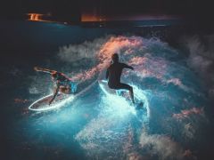 zwei Surfer mit leuchtenden Surfboards surfen zusammen auf der Welle während der Pure Surf Night in der Jochen Schweizer Arena