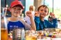 ThumbnailKindergeburtstag in der Jochen Schweizer Arena: Zwei Jungen sitzen fröhlich an einem bunt gedeckten Tisch mit Deko und Getränken und zeigen lachend Handzeichen in die Kamera.